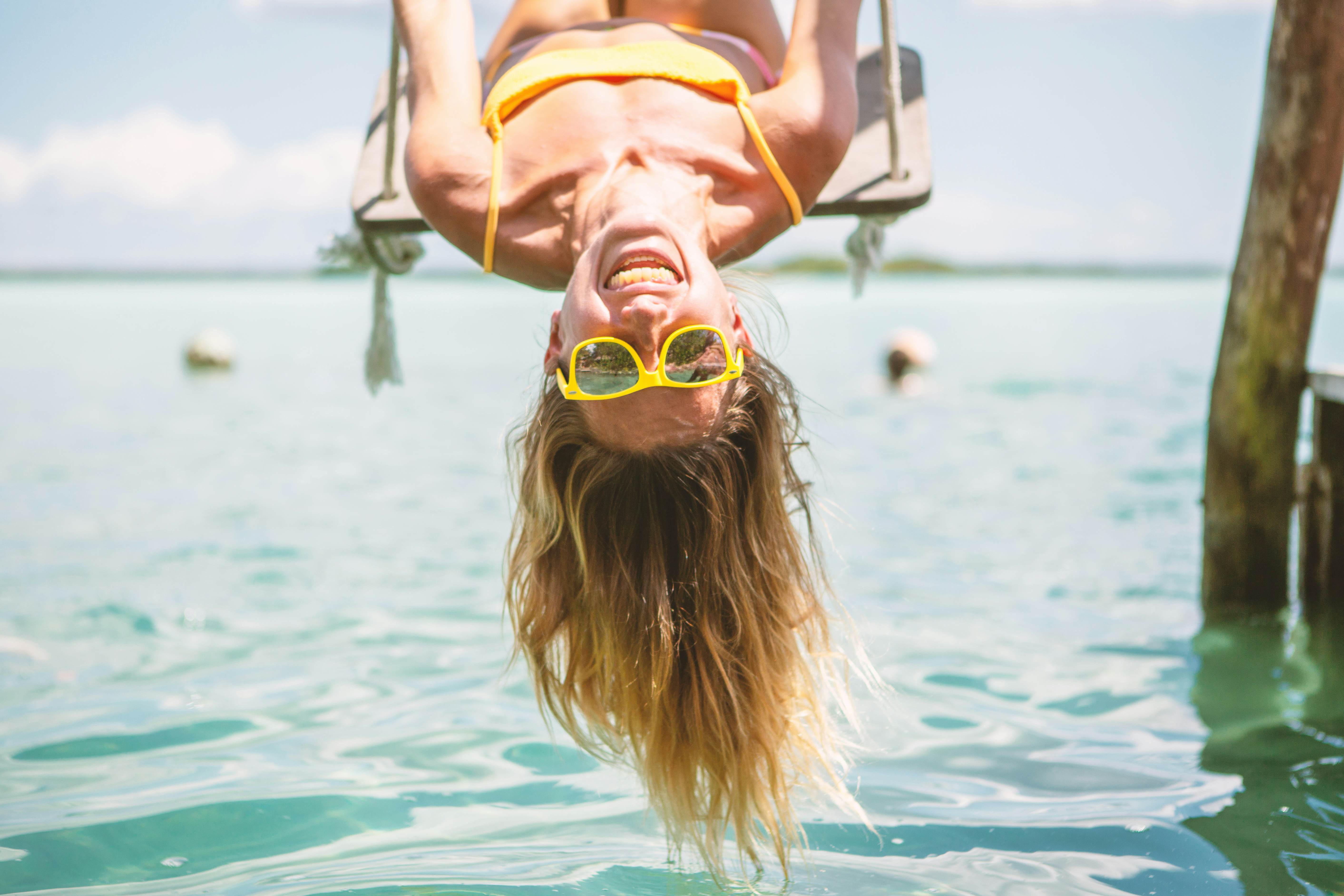Woman playing on swing over the sea in Mexico, she is wearing yellow sunglasses that match her yellow bathing suit and she is smiling joyously. 